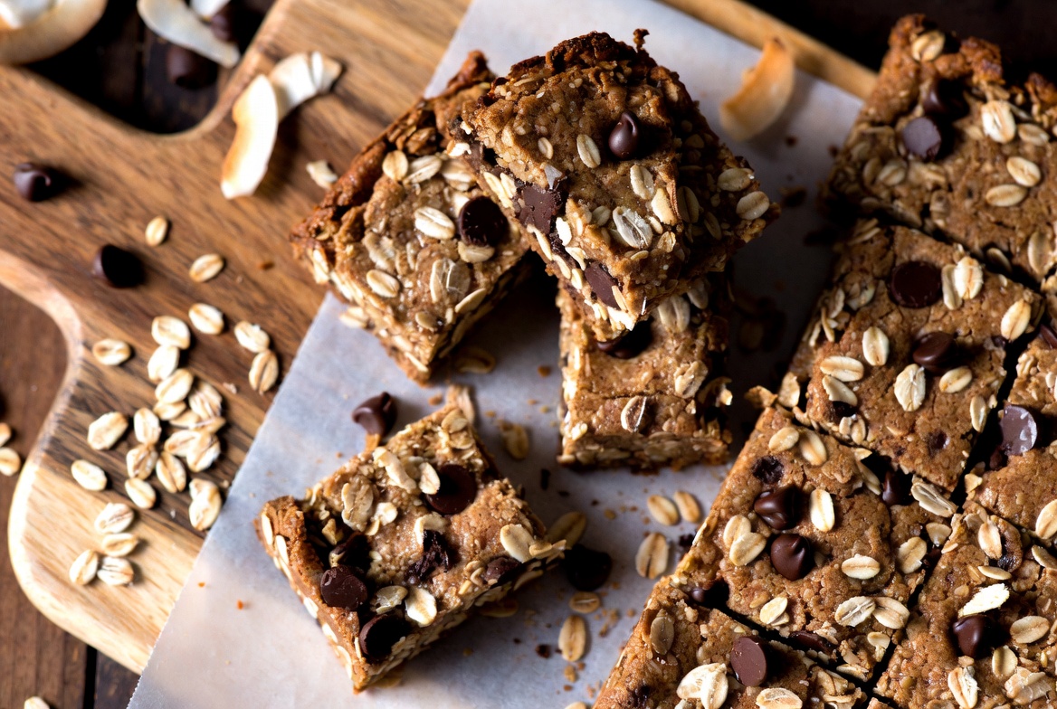 Baked nut butter protein bars cut into squares on parchment, topped with oats and dark chocolate chips
